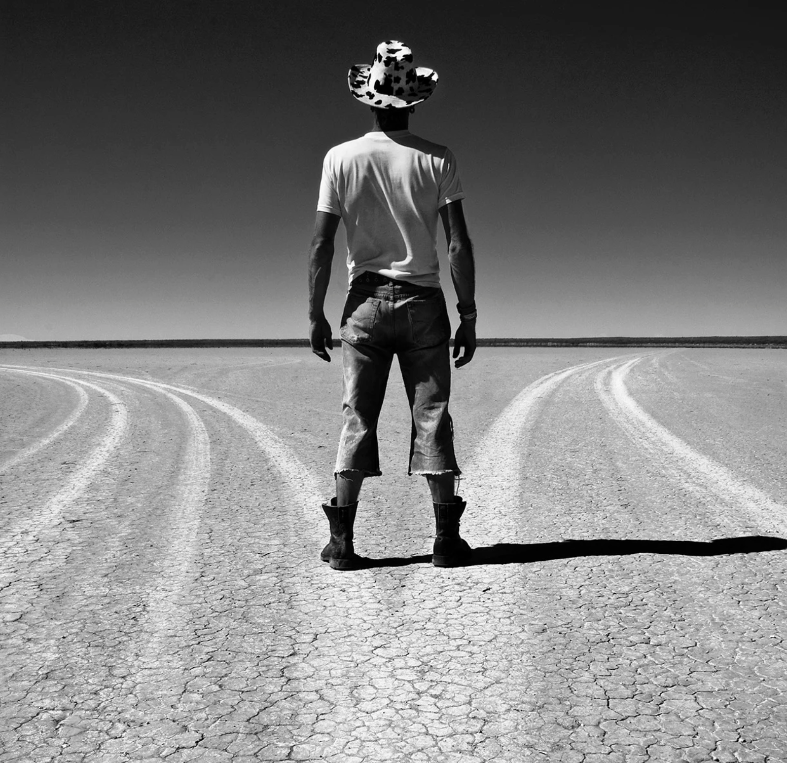 A black and white photograph of a David Ballam standing on cracked ground facing away from the camera, wearing a light-colored t-shirt, dark jeans, boots, and a cow-print hat.