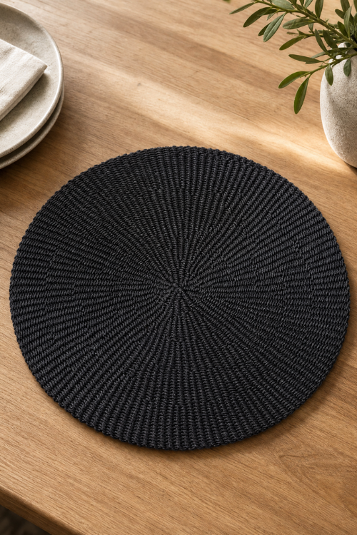 Baba Tree Black woven placemat on a wooden table with a plant and plates in the background
