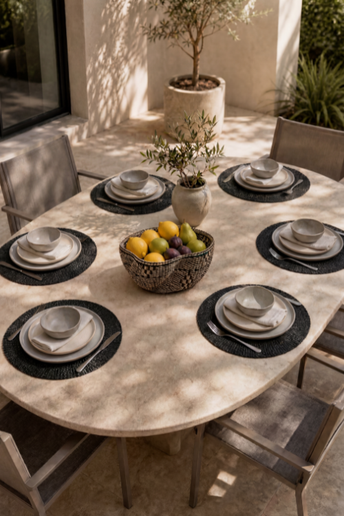 Baba Tree placemats on a Round outdoor dining table set with plates, bowls, and a fruit bowl in a garden setting.