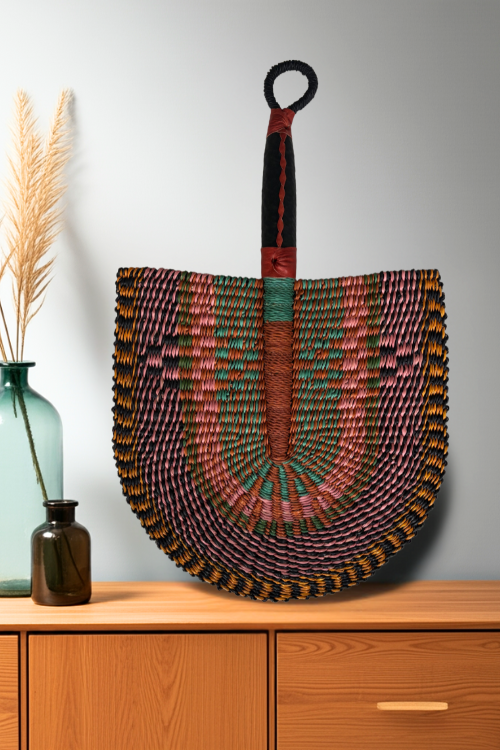 The Baba Tree Handcrafted Bolga Decorative Fan – Vibrant Mirage placed on a console, leaned against a wall.