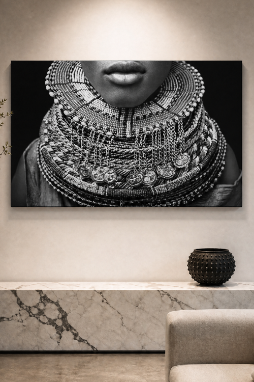 A large black and white canvas African wall art print depicting a close-up of a person wearing an elaborate layered silver necklace, displayed on a beige wall above a marble console table with a textured vase.