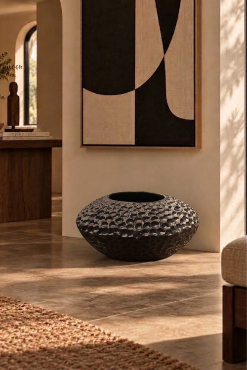 A textured, dark charcoal-colored round planter pot with a raised geometric pattern, displayed in a warm, modern interior setting with a beige wall, abstract art, and stone flooring.
