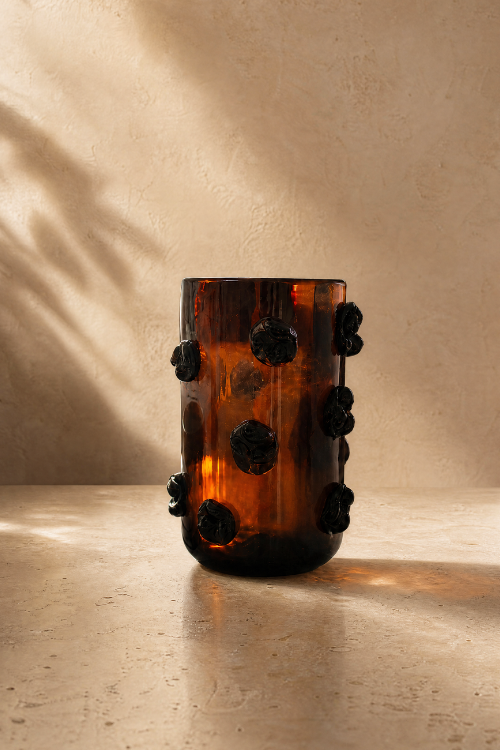 An Nywele vase dark umber with a cylindrical body and raised dark floral accents, photographed on a warm beige surface against a textured beige wall with soft shadow overlays.