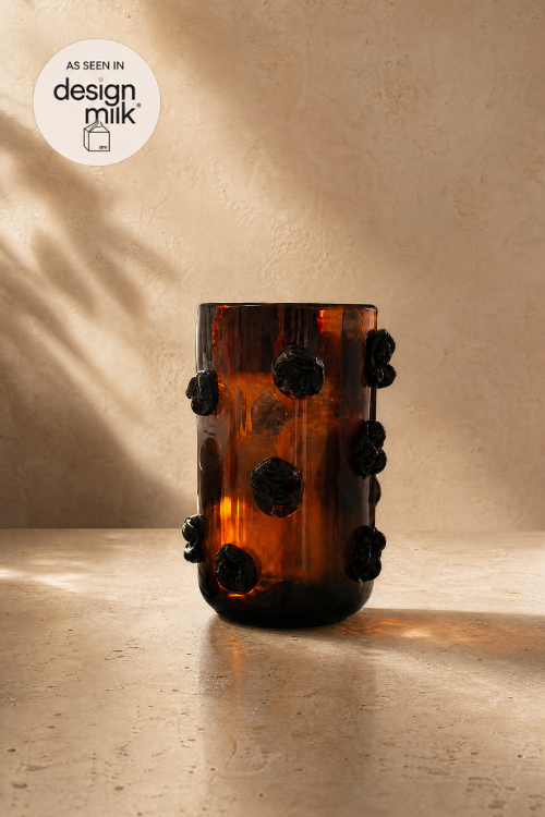 The Nywele vase dark umber in translucent glass vase with dark textured pom-pom accents, set against a warm beige textured wall with soft leaf shadows.