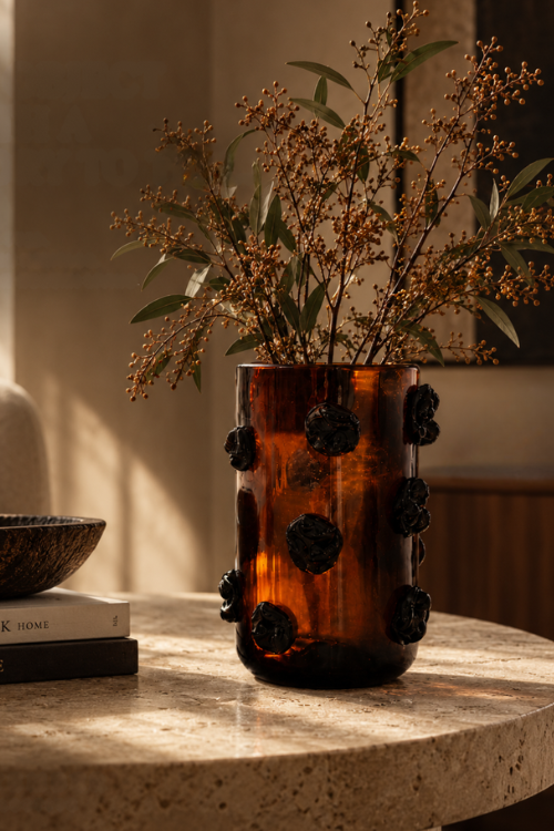 The Nywele dark umber glass vase with dark floral accents sits on a stone tabletop in warm sunlight, holding dried branches with green leaves and small golden-brown buds.