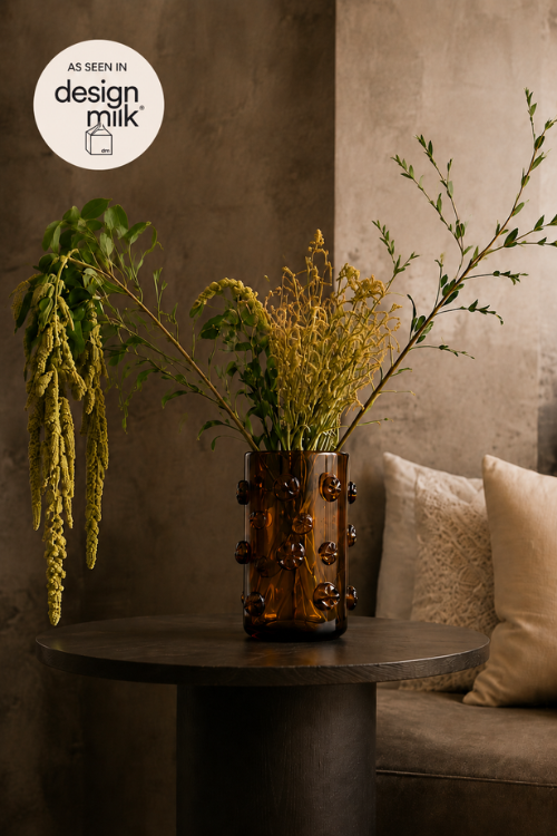 An amber-brown glass Nywele vase with a textured, bubble-like pattern, filled with dried yellow and green botanicals, displayed on a dark round table in a modern living room setting.