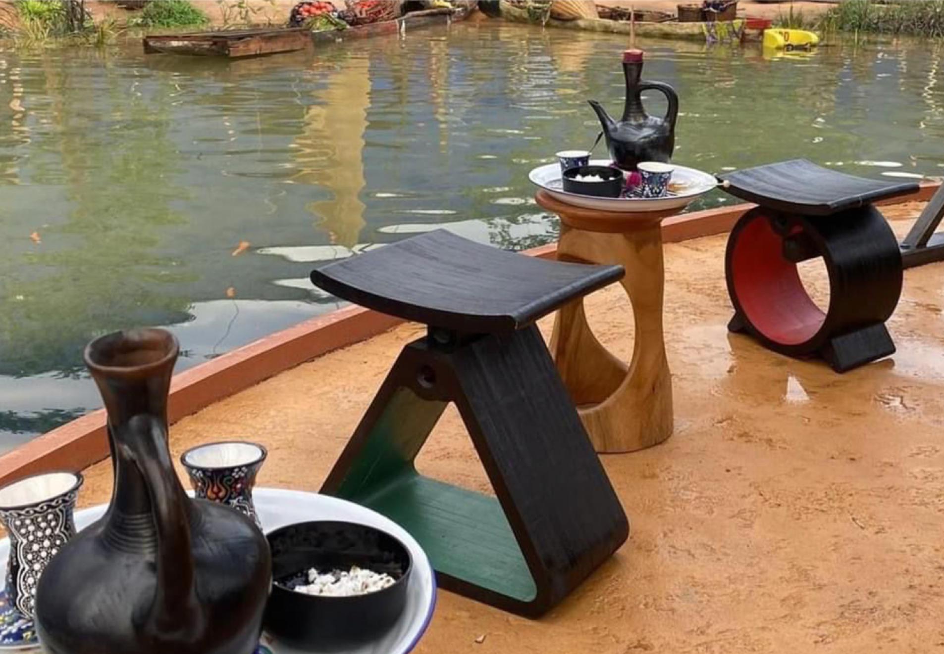 Tekura Wooden stools and side table by a body of water with fishing boats in the background. A scene from the Black panther movie.