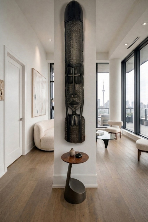A tall, dark carved Obaa african wall sculpture stands on a small round side table in a modern interior hallway with wood flooring, white walls, and large windows.