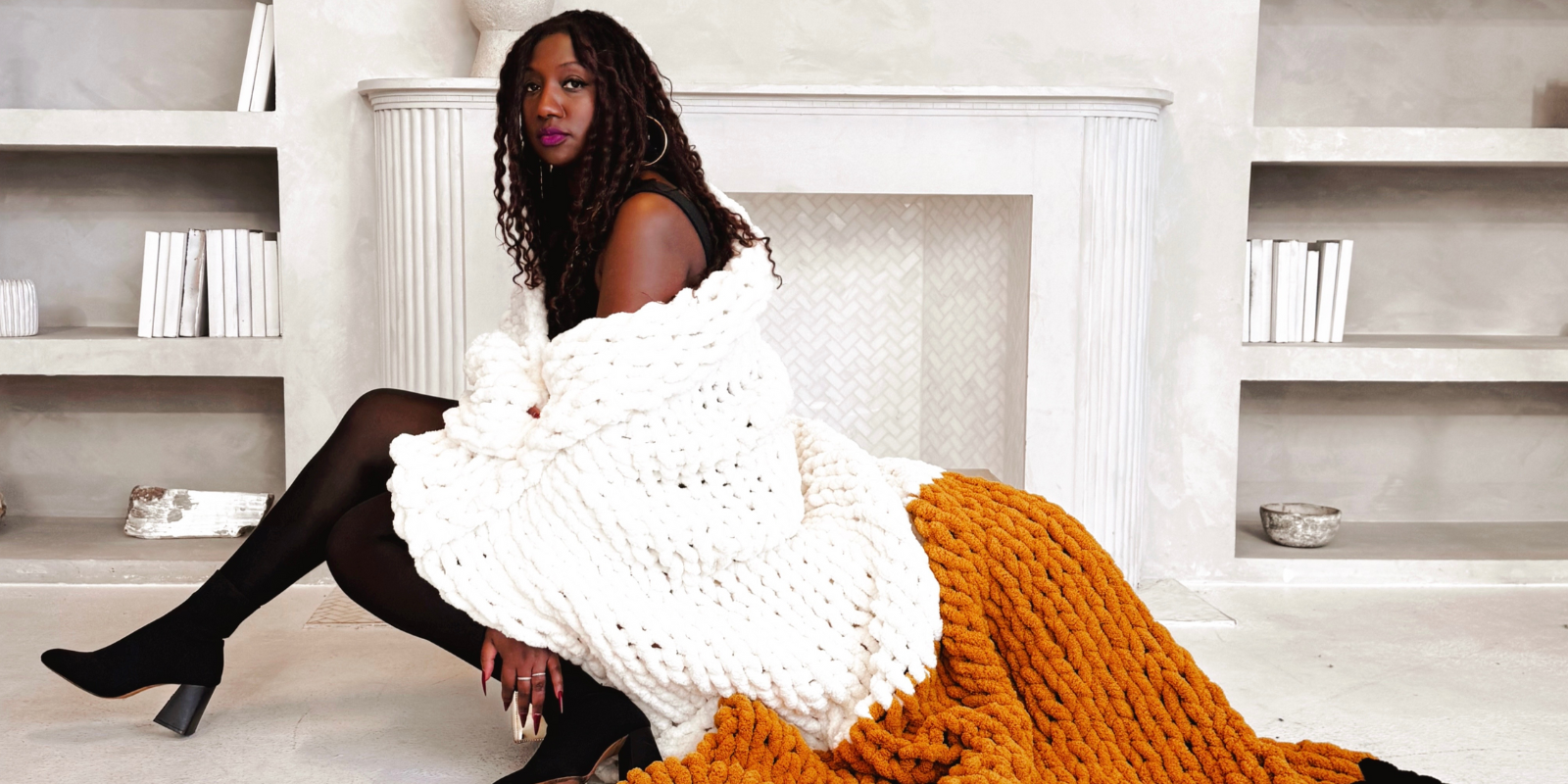 Founder of Villa aburi, Stacey, sitting on a couch with a white and orange blanket in a room with bookshelves
