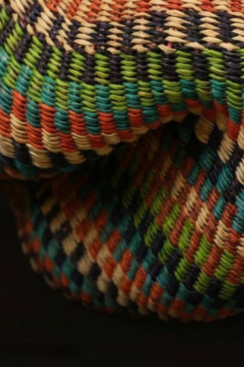 A close-up of a handwoven basket with a colorful geometric pattern of teal, green, orange, beige, and black threads.