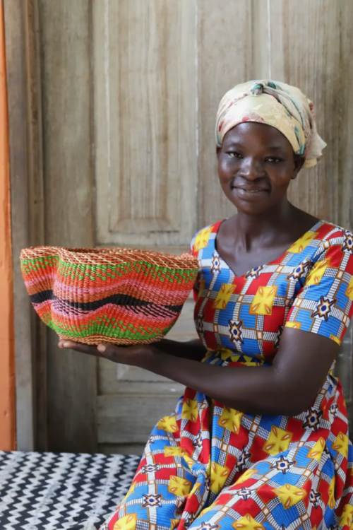 Artisan holding a Baba Tree Petite Pakurigo Wave Basket - Earthy Blossom in pink, orange, green and black in front of a wooden door.