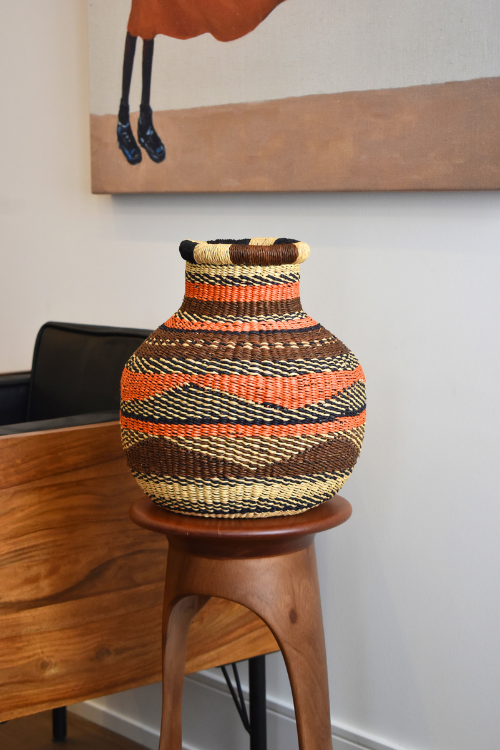 Woven Baba Tree Jemima 10 Cows Tiny Basket with striped pattern on a wooden Djembe side table against a white wall.
