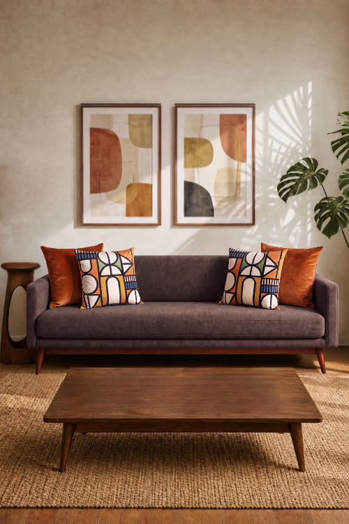 A mid-century modern gray fabric sofa in a styled living room, decorated with orange and patterned throw pillows, set against a textured beige wall with two framed abstract prints and a wooden coffee table.