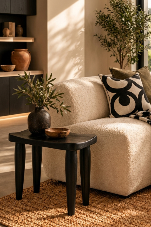 A modern beige boucle sofa in a warm, sunlit living room setting, styled with a black and cream patterned accent pillow, a small black anantu bench side table with a dark vase of greenery, and a woven jute rug.