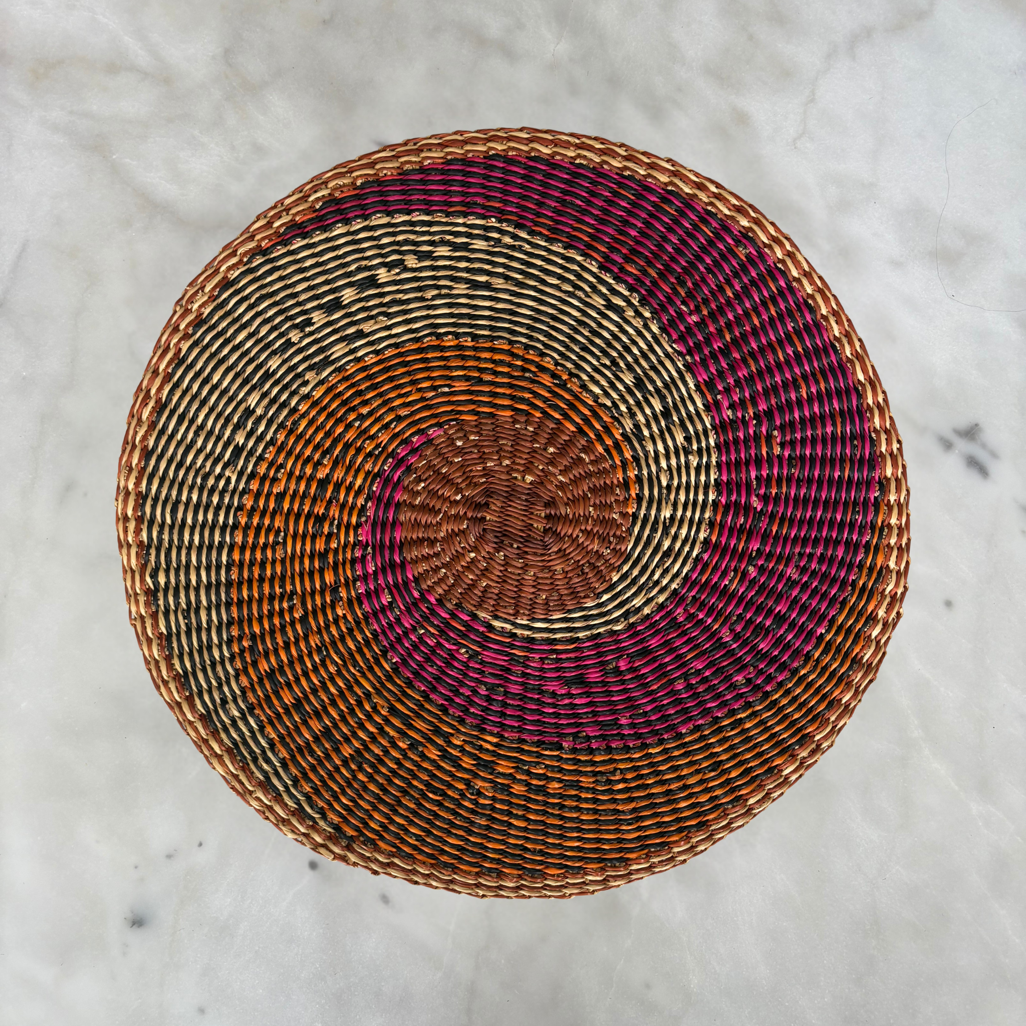 Baba Tree Placemats - African Tide. Woven basket with concentric circle pattern on a marble surface from Baba Tree. Handwoven placemat in pink, orange, natural.