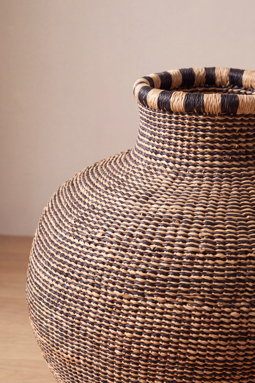 A close-up of Jemima-10-Cows-Basket-Ebony-Natural-Coil with a rounded body and a flared rim, made from natural tan fibers with dark brown to black woven accents, set against a neutral beige background.