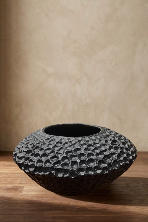 A dark textured ceramic Mdomo vessel  with a hammered, sculptured-like surface finish, displayed on a wooden surface against a neutral beige wall.