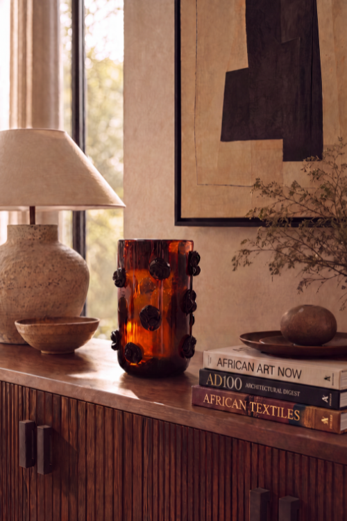 Nywele vase dark umber with dark floral accents sits on a wooden console table in a styled interior setting next to a lamp and books.