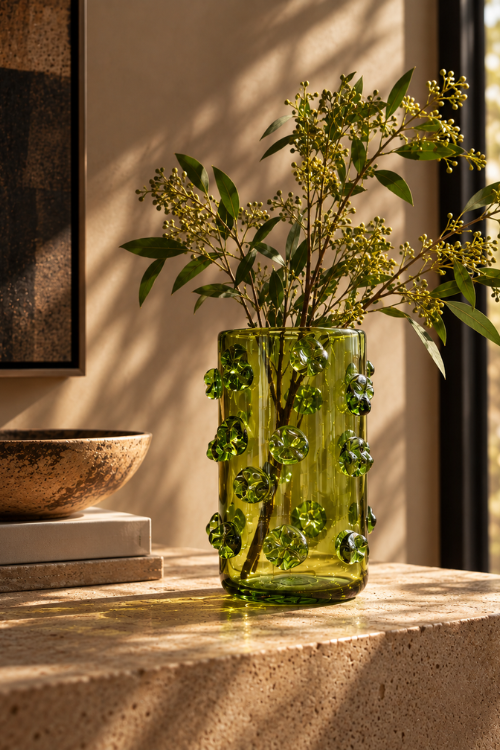 The Nywele vase umber - green glass vase with raised bubble details, filled with green leaves and small yellow flowers, styled on a stone surface in warm sunlight.