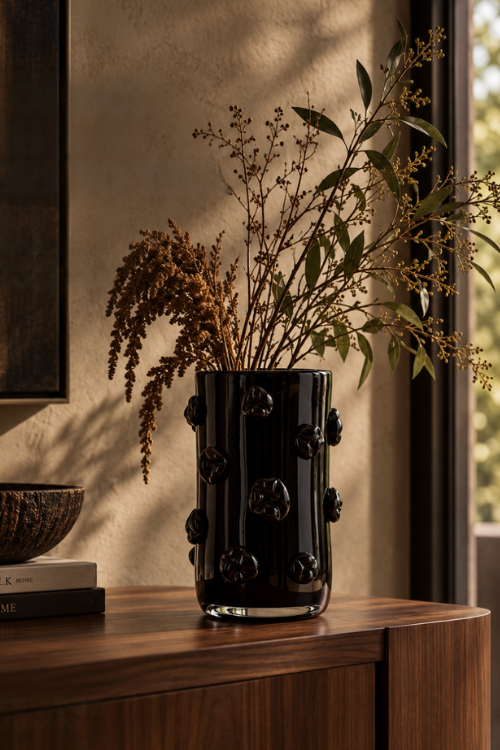 The Onyx black nywele glass vase with raised floral details, filled with dried botanical stems and green leaves, displayed on a wooden console table in a warmly lit interior setting.