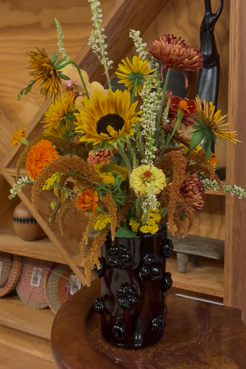 A rustic floral arrangement featuring sunflowers, orange mums, and dried ornamental grasses in a dark brown glossy, Nywele vase dark umber, displayed on a wooden pedestal in front of a wooden shelving unit.