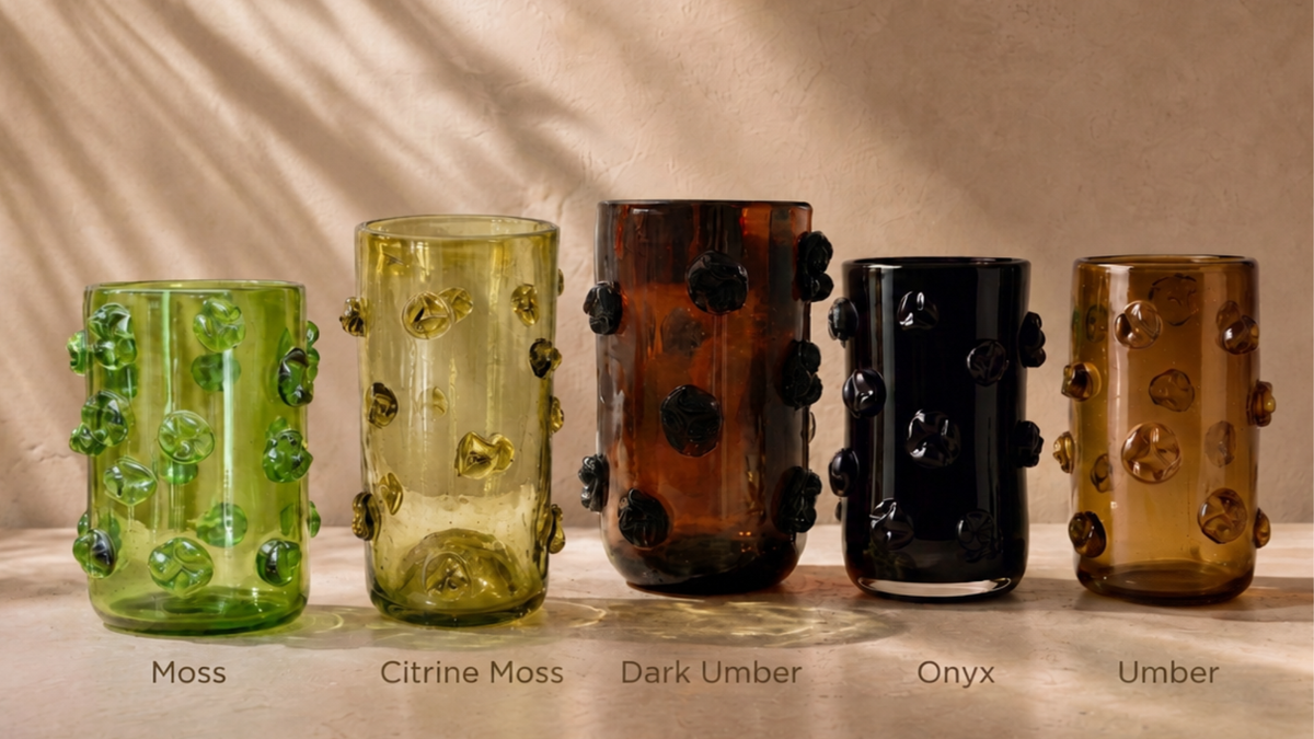Five textured nywele glass vases with bubble-like raised details are lined up on a beige fabric surface. From left to right, the colors are Moss green, Citrine Moss yellow-green, Dark Amber brown, Onyx black, and Amber brown.