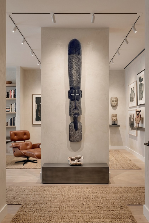 A tall, dark carved African wall sculpture mounted on a beige pillar in a modern gallery or showroom interior with track lighting, framed art, and a brown lounge chair visible in the background.