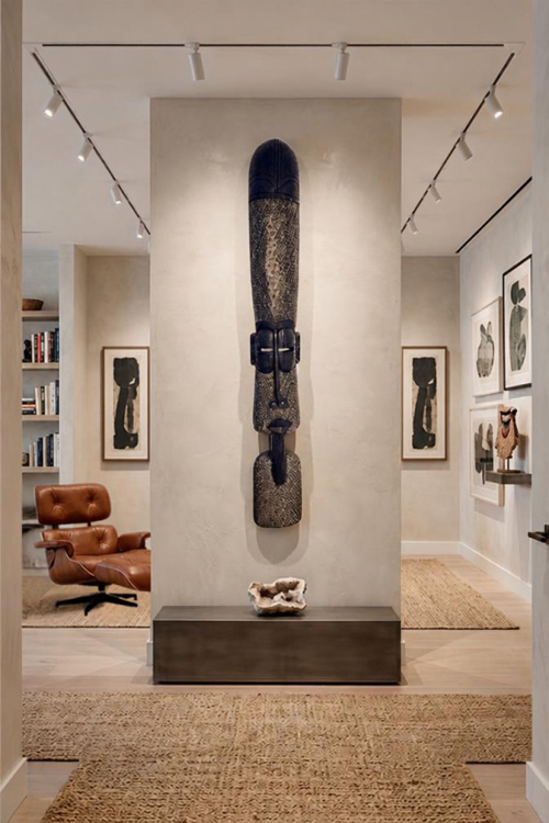 A tall, dark carved African wall sculpture mounted on a central beige pillar in a modern, well-lit gallery or showroom interior with wood floors, woven rugs, and framed art.