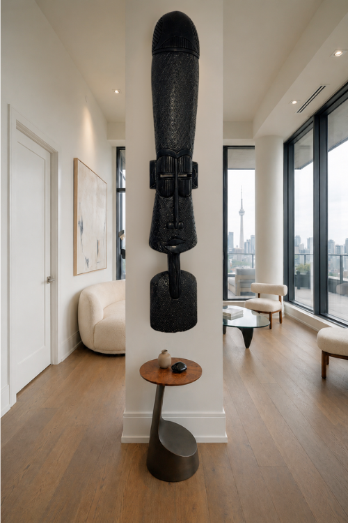 A tall, black textured Obarima African wall totem art piece displayed on a white pillar in a modern, sunlit living room with hardwood floors and large windows.
