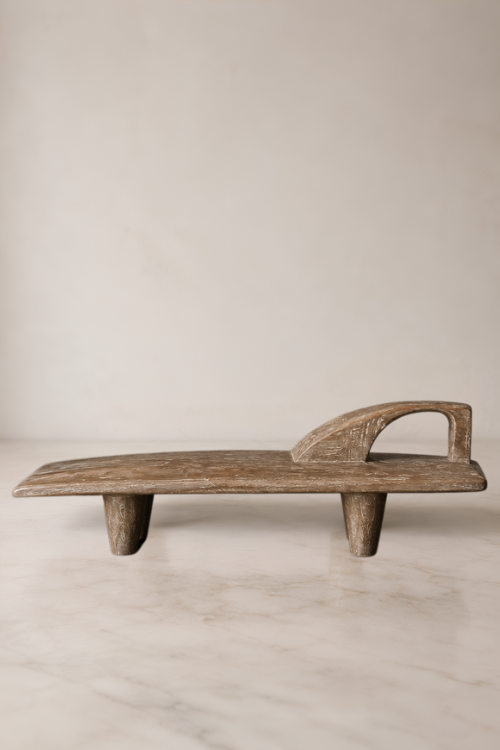 A Senufo-Sculpture-Stand-by-Tekura-with a weathered brown finish, featuring a curved backrest and four short legs, set against a neutral beige background.