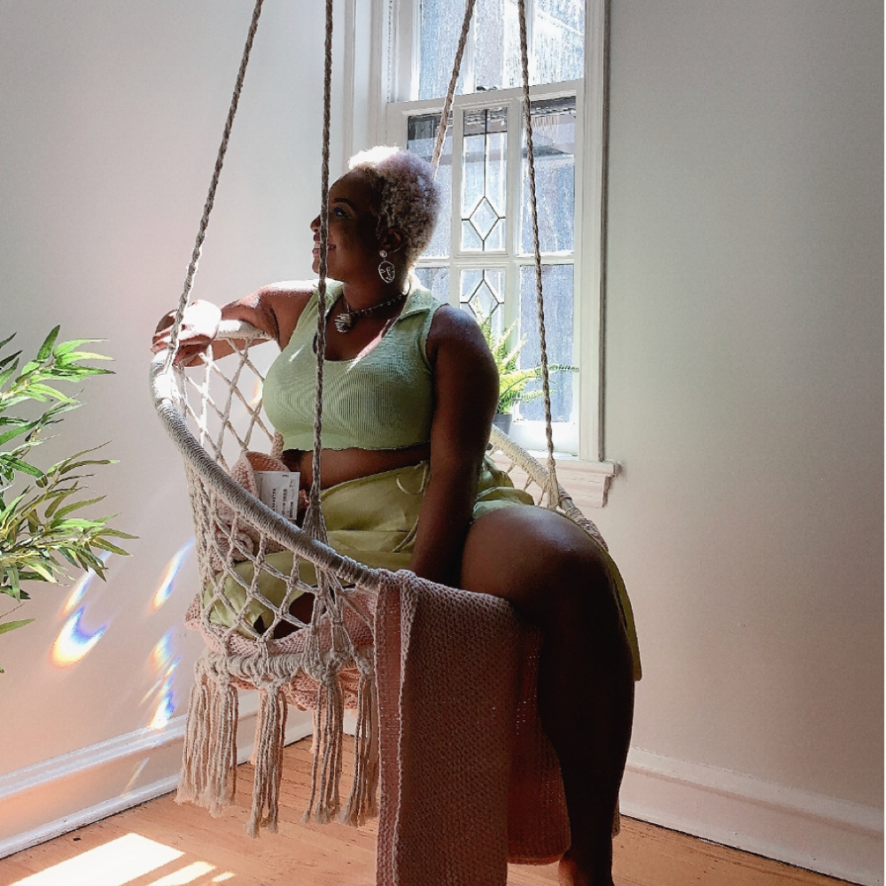 Tamara Tashna sitting in a macrame swing in a room with a window and plant.