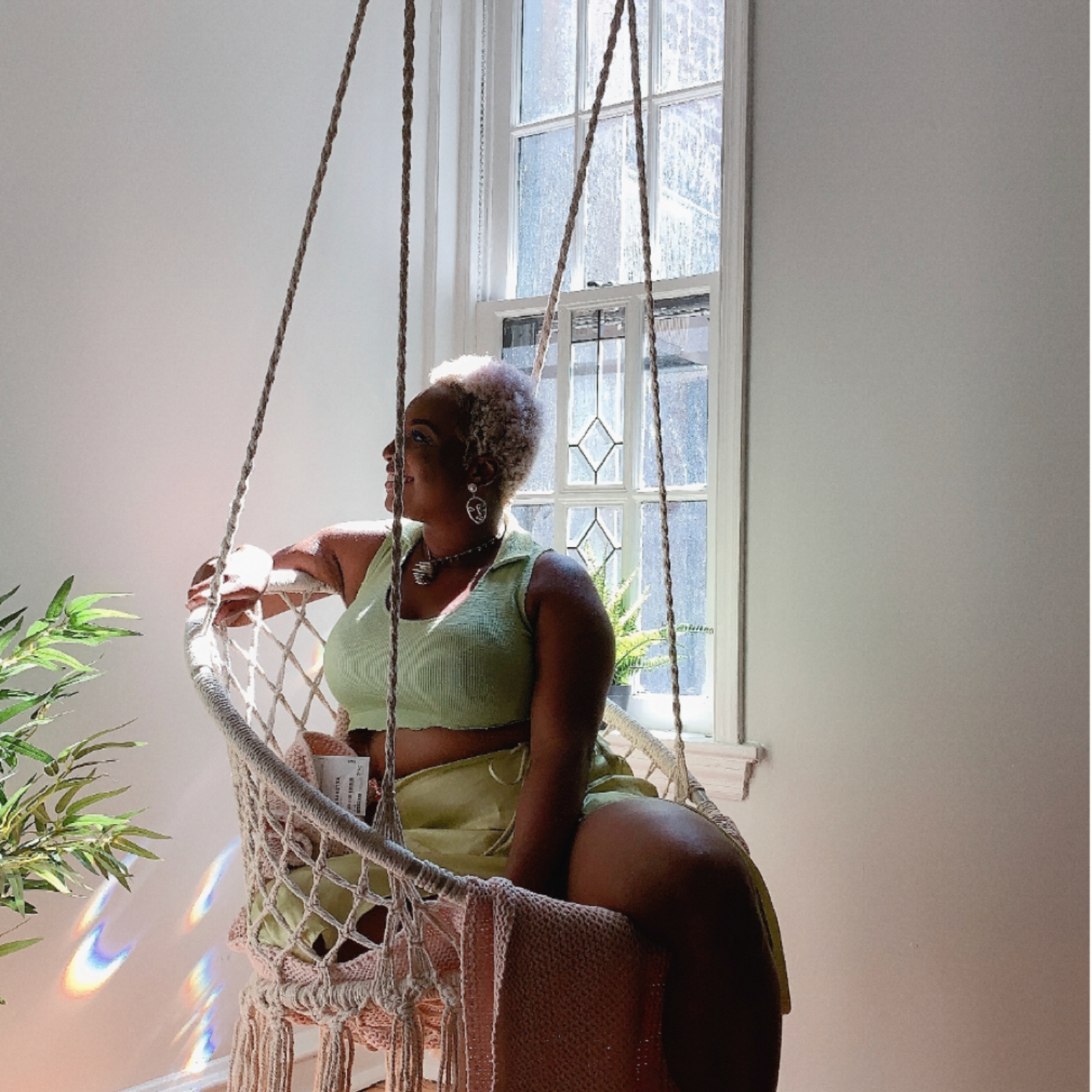 Tamara Tashna sitting in a macrame swing by a window with plants around.