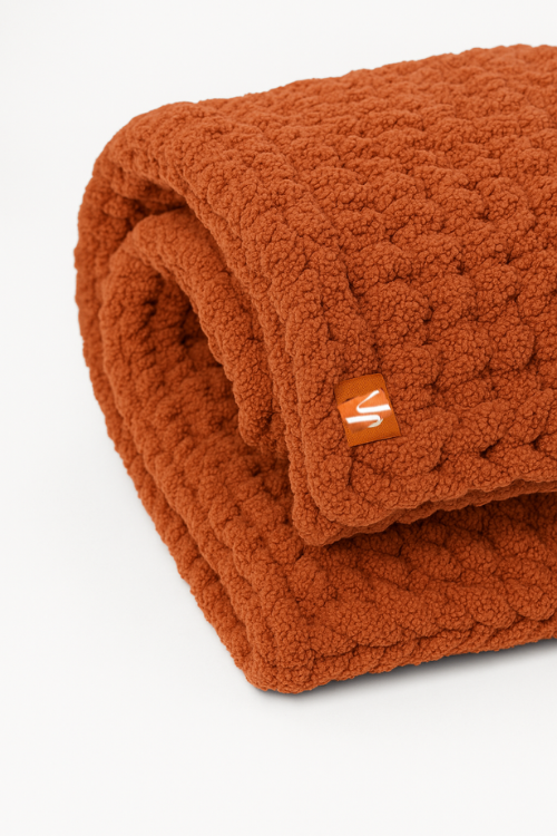 Folded burnt orange hand knitted throw  with a brand logo on a white background
