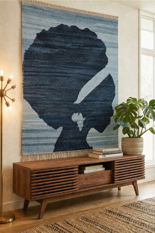 A large woven wall hanging with a stylized black silhouette over blue and gray horizontal stripes, displayed above a mid-century modern wooden console in a bright living room with a plant and lamp.