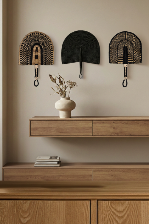 Three decorative bolga fans on a wall above a wooden console table with a vase and books.