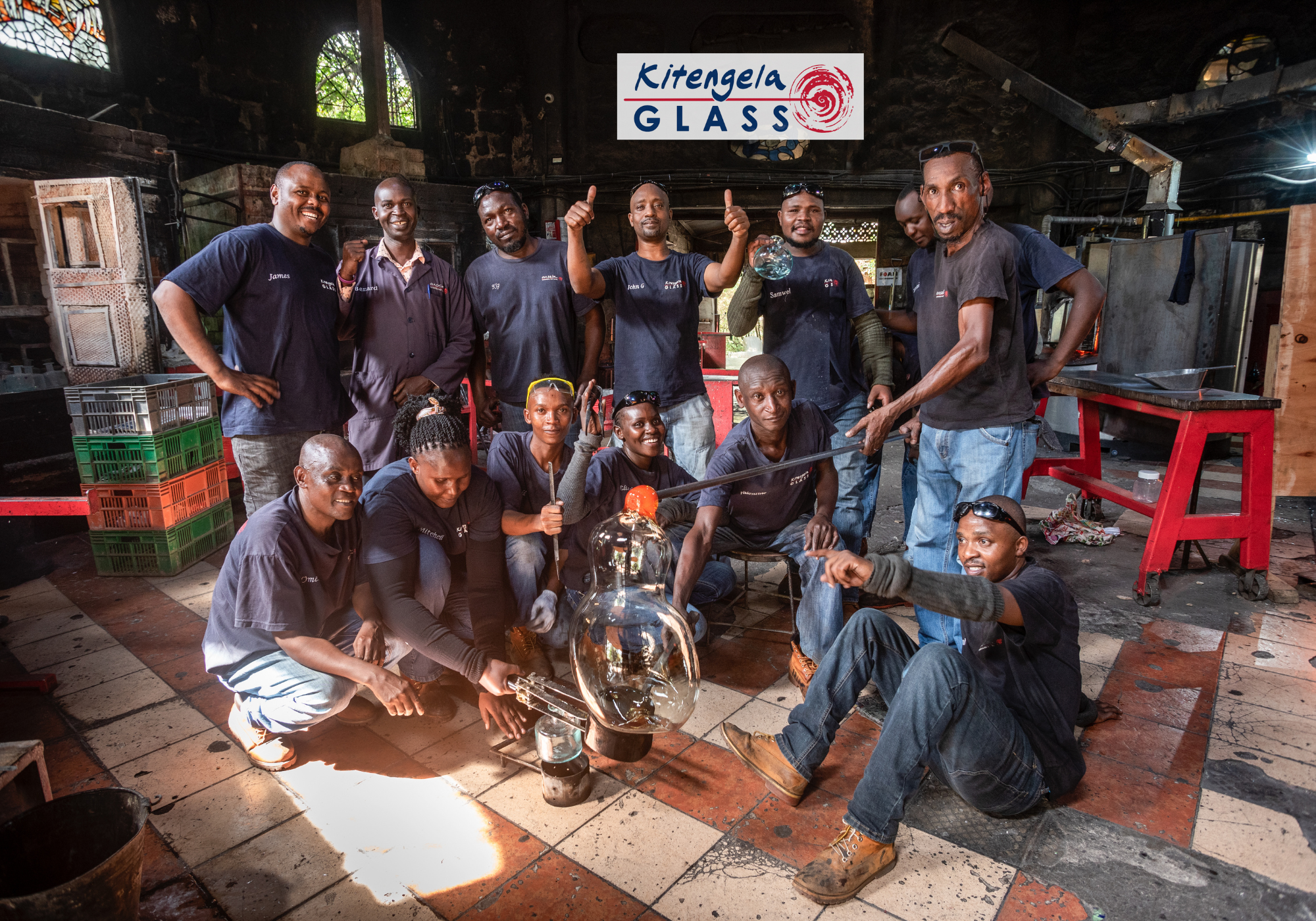 Group of artisan posing with a large glass object in the kitengela glass workshop setting, Kitenega Glass logo visible.