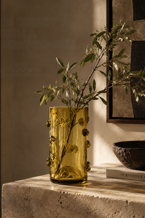 The nywele citrine moss vase with a textured, bubble-like pattern sits on a stone table in a warm, sunlit room next to a dark bowl and a book